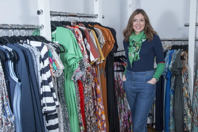 Commerçante souriante dans sa boutique de vêtements, entourée de portants de robes colorées