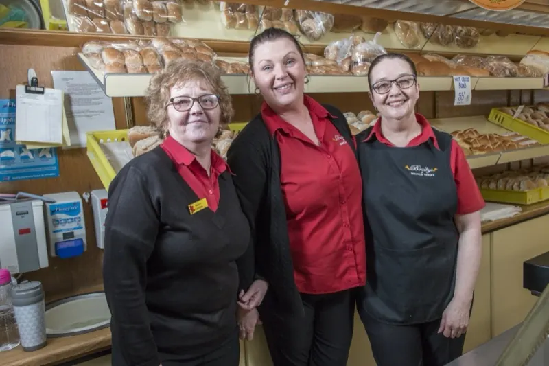 Trois employées souriantes posant ensemble dans leur boulangerie, devant les étagères de pains