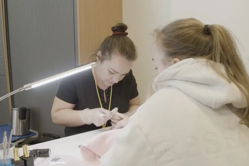 Prothésiste ongulaire gantée réalisant une manucure sur une cliente, sous une lampe de travail