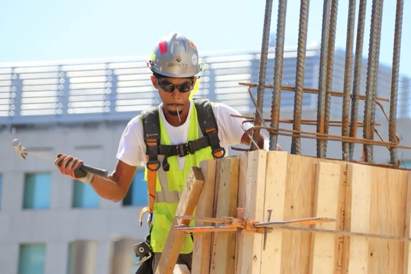 Ouvrier du bâtiment équipé d'un casque, de lunettes, de gants et d'un harnais, montant un coffrage de poteau sur un chantier